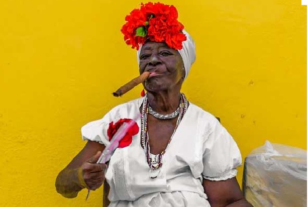 Person in traditional white attire with red flowers and cigar, seated against a bright yellow wall.