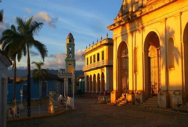 Colonial town square with yellow and blue buildings, palm trees, and a clock tower at sunset.