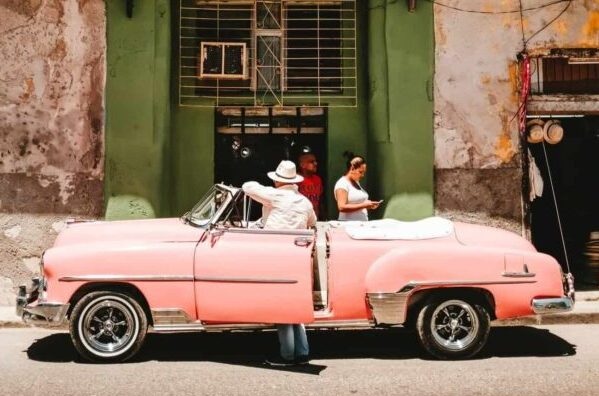 "Vintage pink convertible parked on a street with four people around it, in front of a weathered green and beige building with peeling paint and barred windows."