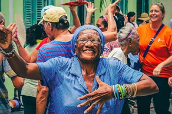 "Older woman in blue shirt and headscarf dancing joyfully with a group of people at a festive outdoor event."